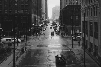 mezzalyn. Blog. Uma paisagem urbana em preto e branco mostra uma rua molhada e vazia ladeada por prédios altos. Alguns pedestres espalhados atravessam a rua, enquanto carros estão estacionados e se movendo ao lado. Céus nublados adicionam uma atmosfera melancólica à cena urbana.