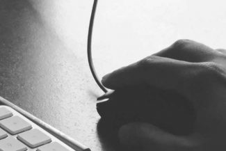 General Terms and Conditions (GTC) Uma imagem em preto e branco da mão de uma pessoa usando um mouse com fio ao lado de um teclado em uma mesa escura. A cena é minimalista, com luz natural suave vindo da janela.