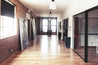 Privacy Policy Um corredor de escritório moderno e iluminado com pisos de madeira, paredes de tijolos e portas de vidro. Pinturas estão apoiadas na parede esquerda, e uma bicicleta é visível perto de uma grande janela no final do corredor.