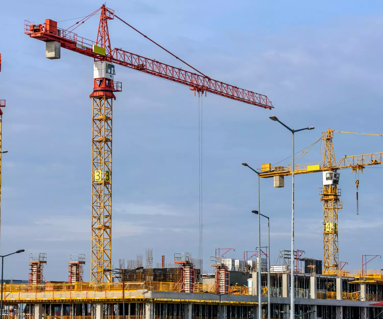 Um canteiro de obras com vários guindastes elevando-se sobre um edifício parcialmente construído promete um futuro de habitação saudável. Os guindastes estão contra um céu nublado, e o edifício é cercado por andaimes. Postes de luz são visíveis no primeiro plano, iluminando o progresso.