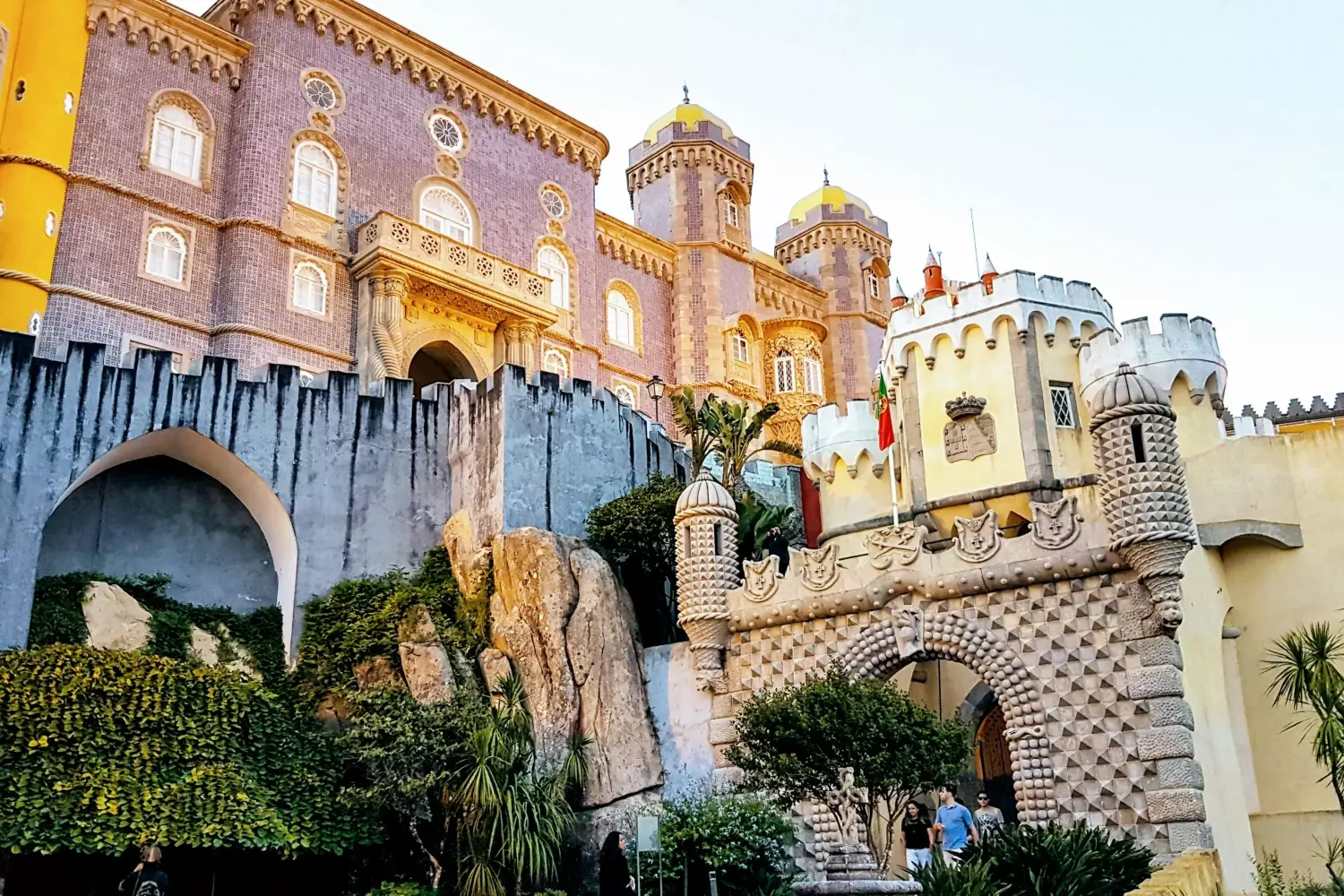 A imagem mostra a fachada colorida e ornamentada do Palácio da Pena em Sintra, Portugal. Esta habitação saudável apresenta torres vibrantes amarelas e vermelhas, detalhes arquitetônicos intrincados e vegetação exuberante em primeiro plano, incorporando uma sensação de pureza e beleza natural.