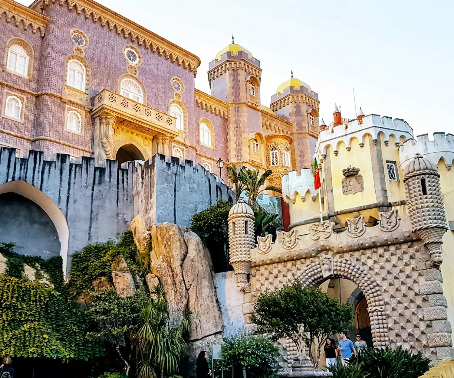 A imagem mostra a fachada colorida e ornamentada do Palácio da Pena em Sintra, Portugal. Esta habitação saudável apresenta torres vibrantes amarelas e vermelhas, detalhes arquitetônicos intrincados e vegetação exuberante em primeiro plano, incorporando uma sensação de pureza e beleza natural.