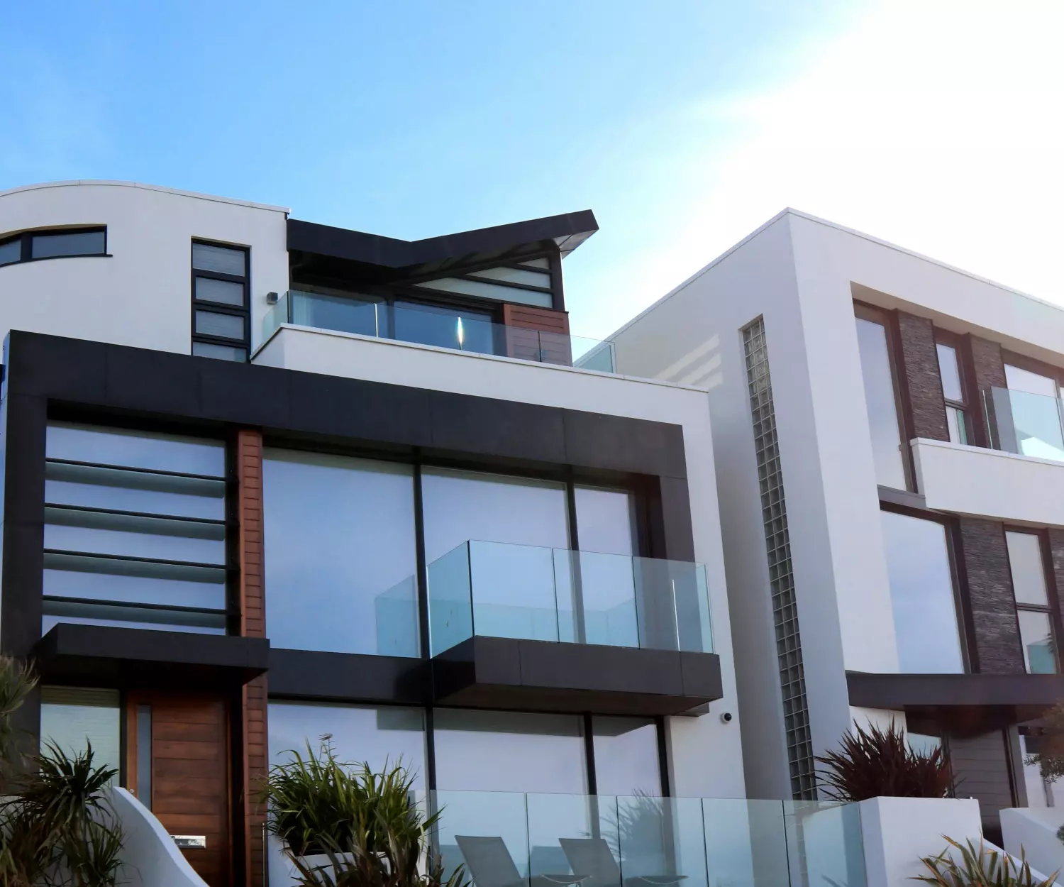 Um edifício moderno de vários andares com arquitetura elegante, com grandes sacadas e janelas de vidro. A fachada combina paredes brancas com detalhes escuros, incorporando a essência da moradia livre de desperdício. Um céu azul claro e ensolarado enfeita o fundo, enquanto palmeiras adornam o primeiro plano.