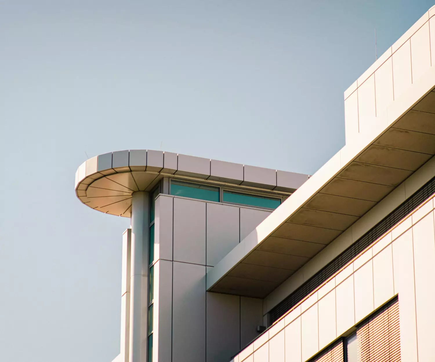 Um edifício moderno com arquitetura angular e elegante e uma extensão cilíndrica ostenta grandes janelas contra um céu azul claro. A luz do sol projeta sombras suaves em sua fachada branca, destacando seu design contemporâneo e livre de toxinas para uma vida mais saudável.