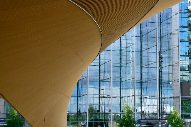 Investing in Sustainability Vista interna de um edifício arquitetônico moderno com um amplo teto de madeira e grandes janelas de vidro, incorporando os princípios da habitação saudável. Reflexos de árvores e nuvens são visíveis no vidro. Algumas pessoas caminham abaixo, dando uma sensação de escala.