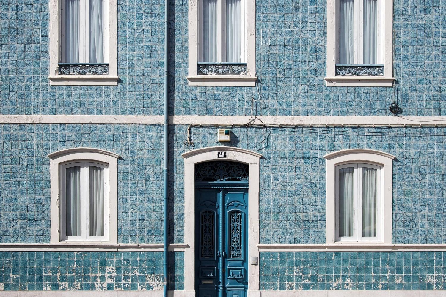 Uma fachada de edifício de azulejos azuis em Portugal exibe três janelas com moldura branca acima de uma única porta azul em arco. O exterior ostenta padrões decorativos, realçados por um poste de iluminação pública na frente. Esta cena pitoresca sutilmente acena para o charme sustentável da nação e as raízes da Revolução Verde. Sustainable: COLOGNE PROJECT 1