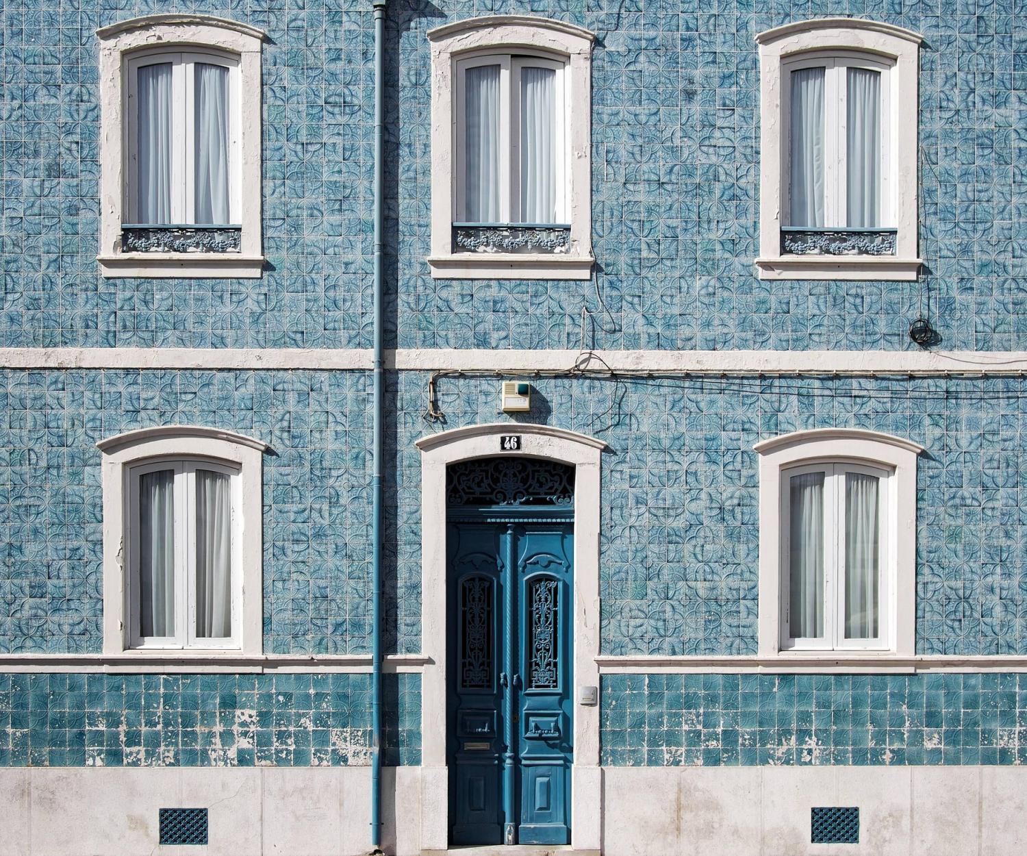 Uma fachada de edifício de azulejos azuis em Portugal exibe três janelas com moldura branca acima de uma única porta azul em arco. O exterior ostenta padrões decorativos, realçados por um poste de iluminação pública na frente. Esta cena pitoresca sutilmente acena para o charme sustentável da nação e as raízes da Revolução Verde. Sustainable: COLOGNE PROJECT 1