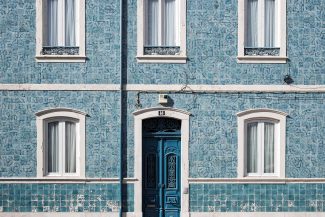 Sustainable? Portugal's Green Revolution: Your Path to a Sustainable Living Dream Uma fachada de edifício de azulejos azuis em Portugal exibe três janelas com moldura branca acima de uma única porta azul em arco. O exterior ostenta padrões decorativos, realçados por um poste de iluminação pública na frente. Esta cena pitoresca sutilmente acena para o charme sustentável da nação e as raízes da Revolução Verde. Sustainable: COLOGNE PROJECT 1